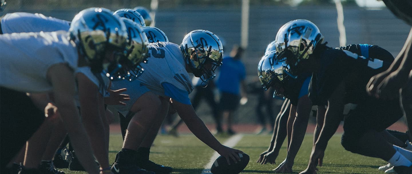 Low-angle shot of offensive and defensive football lines facing each other at the line of scrimmage, with the center's hand on the football, all players wearing reflective chrome helmets.